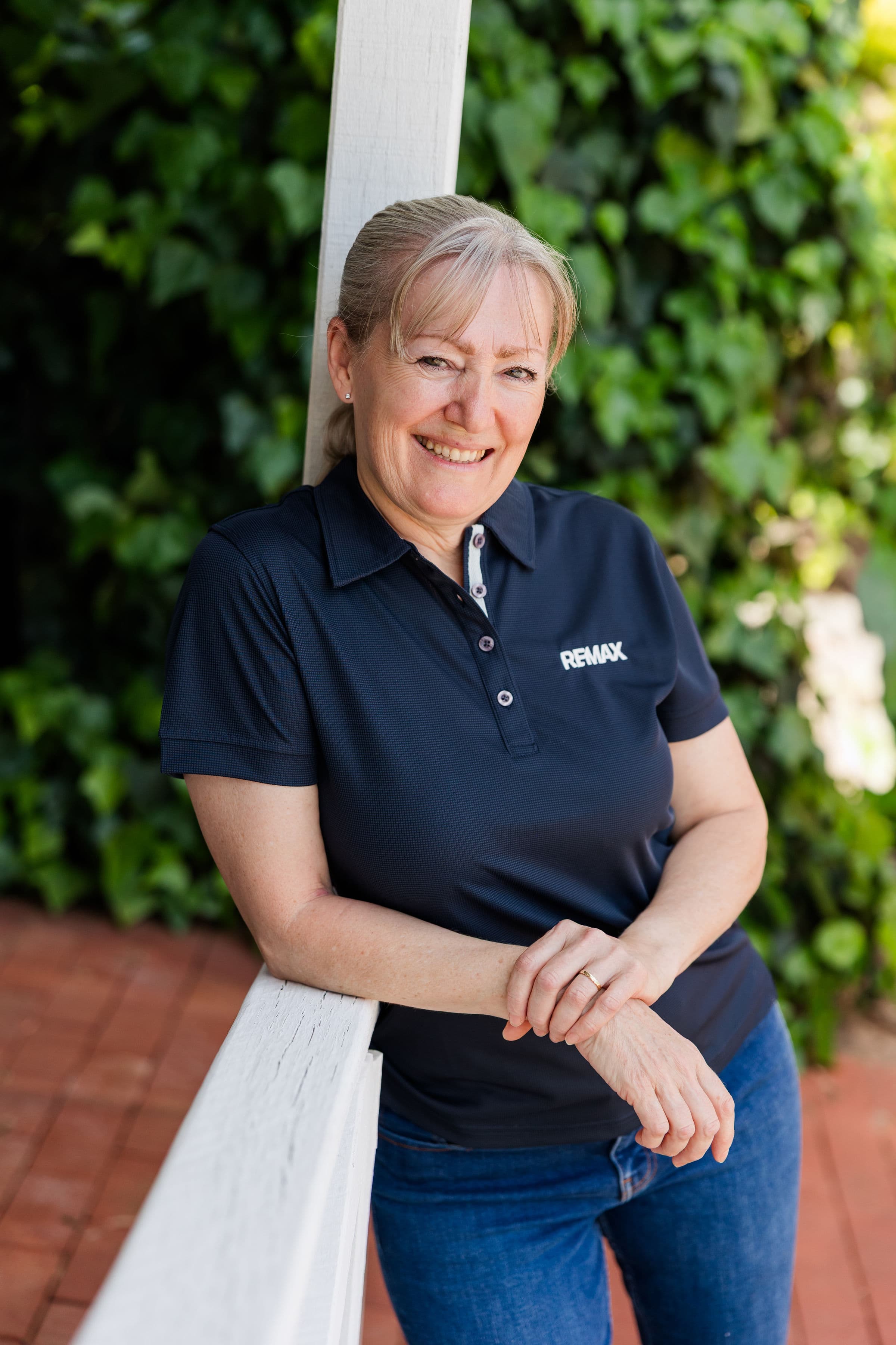 Smiling woman in a navy RE/MAX polo shirt leaning against a white railing outdoors.