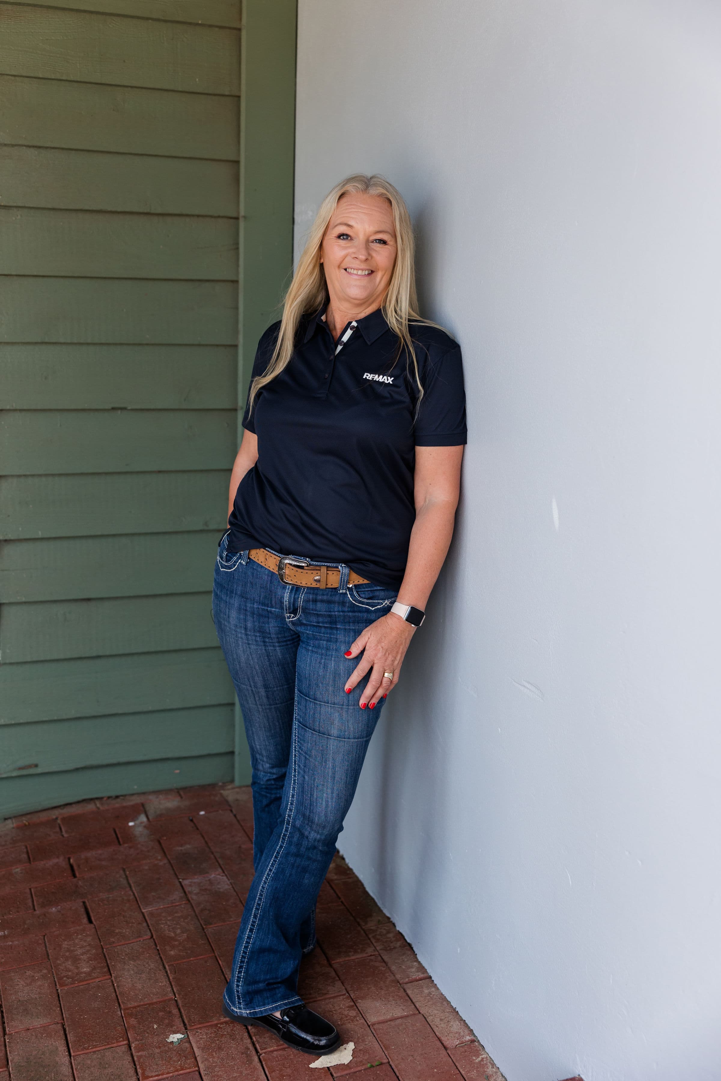 Smiling blonde woman in a navy RE/MAX polo and jeans, leaning against a white wall.